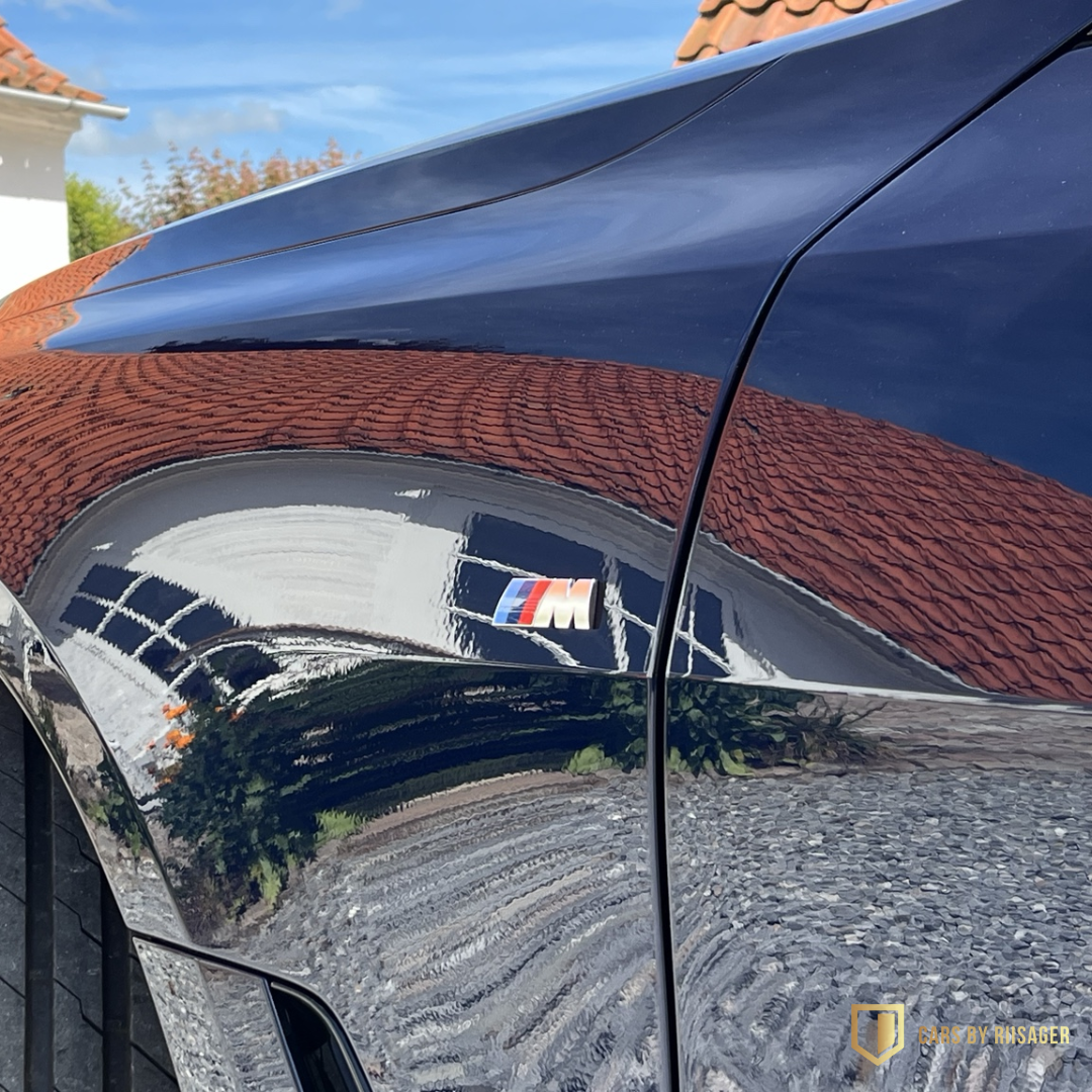 bmw-i4-m-sport-tanzanite-blue-lak Nærbillede af BMW M Sport-logo og Tanzanite Blue Metallic-lak med spejlblank refleksion efter keramisk coating.