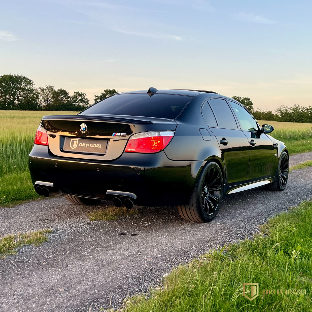 bmw-m5-e60-bagfra-solnedgang BMW M5 E60 set bagfra på landevej ved solnedgang med den ikoniske V10-sedan i profil.